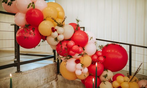 A large balloon garland display, featuring bright pink, orange and pastel-toned balloons, is strung along a stair railing. Red roses and green vines are woven between balloons.