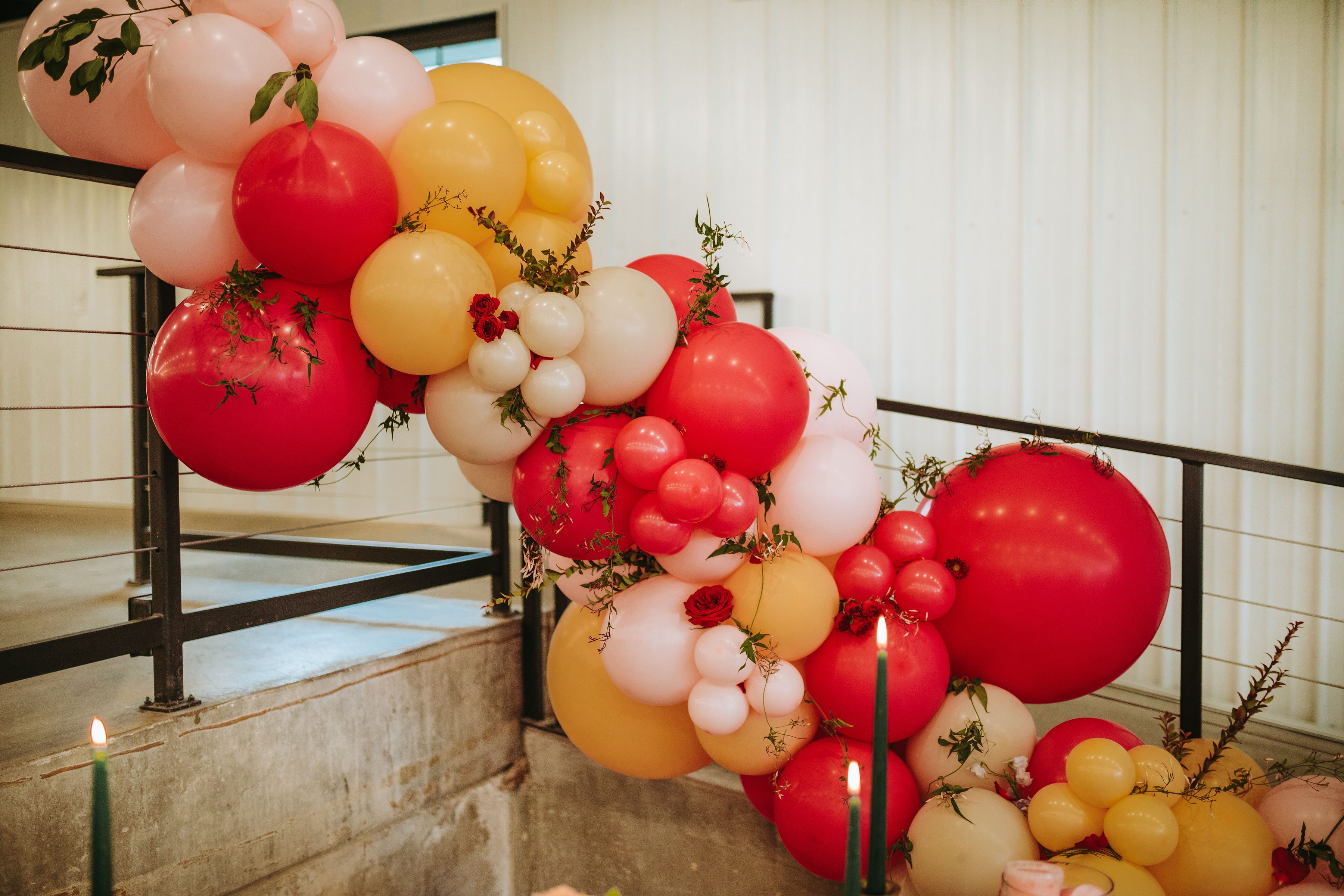 A large balloon garland display, featuring bright pink, orange and pastel-toned balloons, is strung along a stair railing. Red roses and green vines are woven between balloons.