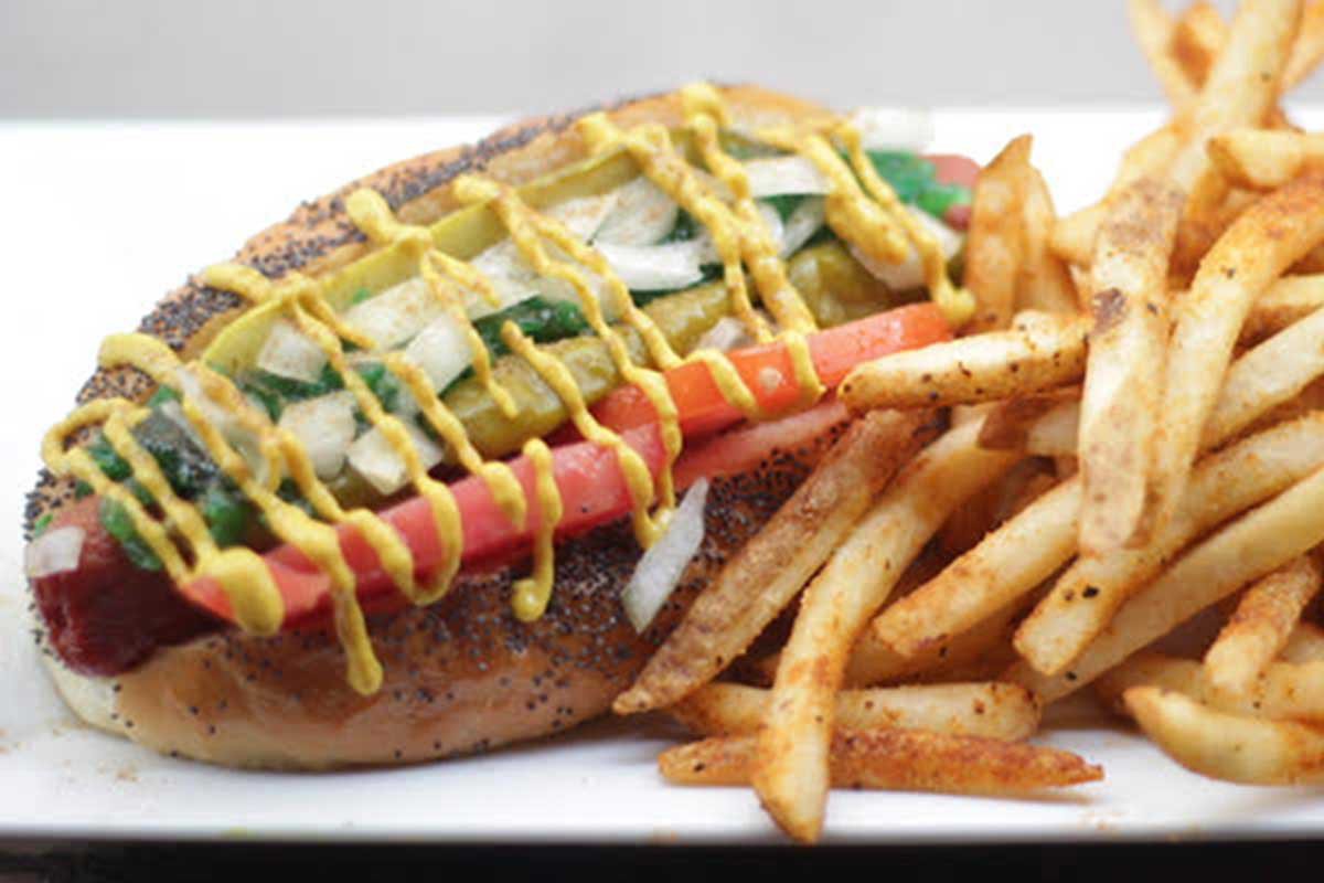 A hot dog covered in veggies and mustard with fries on the side