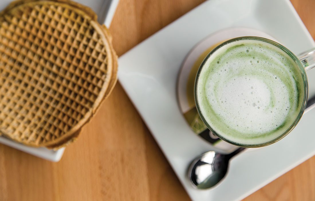 The Amsterdam Fog coffee sits on a white plate with a waffle on the side