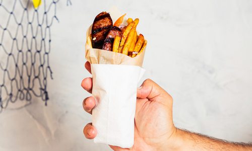 A hand holds up a pita wrap against a white backdrop. Fries, charred sausage and tomato stick out of the the wrap.