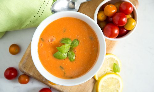 A bowl of gazpacho sits on a wooden slab with baby tomatoes and lemon slices surronding