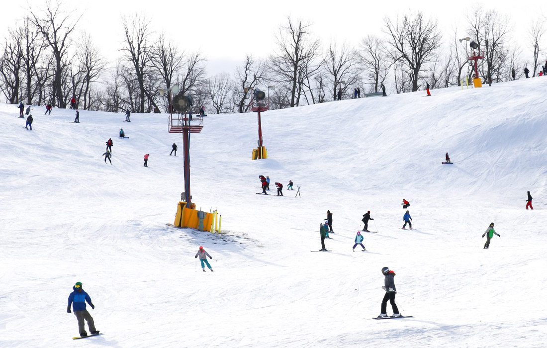 A crowd of people dressed in winter gear ski and snowboard down a snowy hill at Snow Creek Resort.