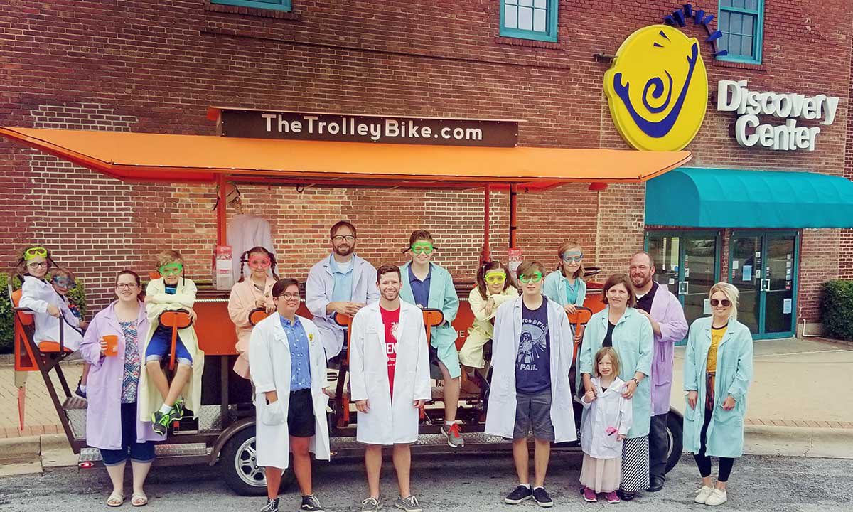 the Trolley Bike staff and Discover Center friends line up in front of the trolley bike parked outside the Discovery Center