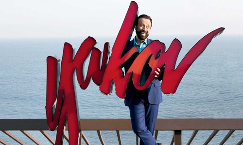 Yakob stands on a pier, water behind him, holding a large sign with his name on it: "Yakov"