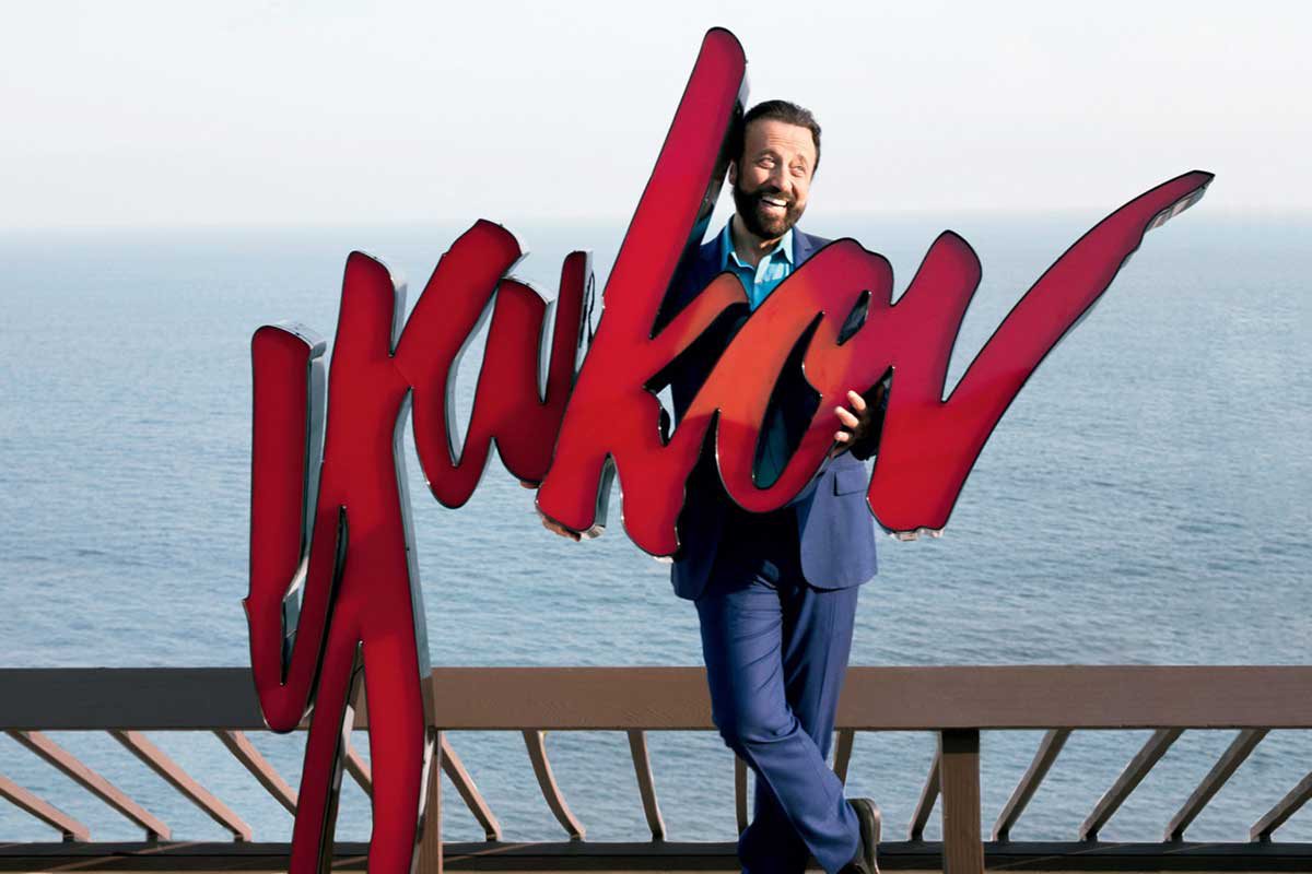 Yakob stands on a pier, water behind him, holding a large sign with his name on it: "Yakov"